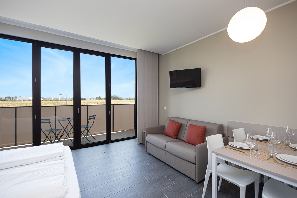 A modern room with a balcony, a sofa, a dining table set for four, and a TV mounted on the wall.