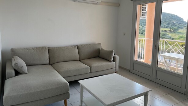 A living room with a grey sectional sofa, a white coffee table, and a sliding glass door leading to an outdoor balcony with a view of greenery and hills. - Très bel Appartement de 71 m², vue sur mer 250 m de la Plage ,du Centre-ville (L'Île-Rousse)