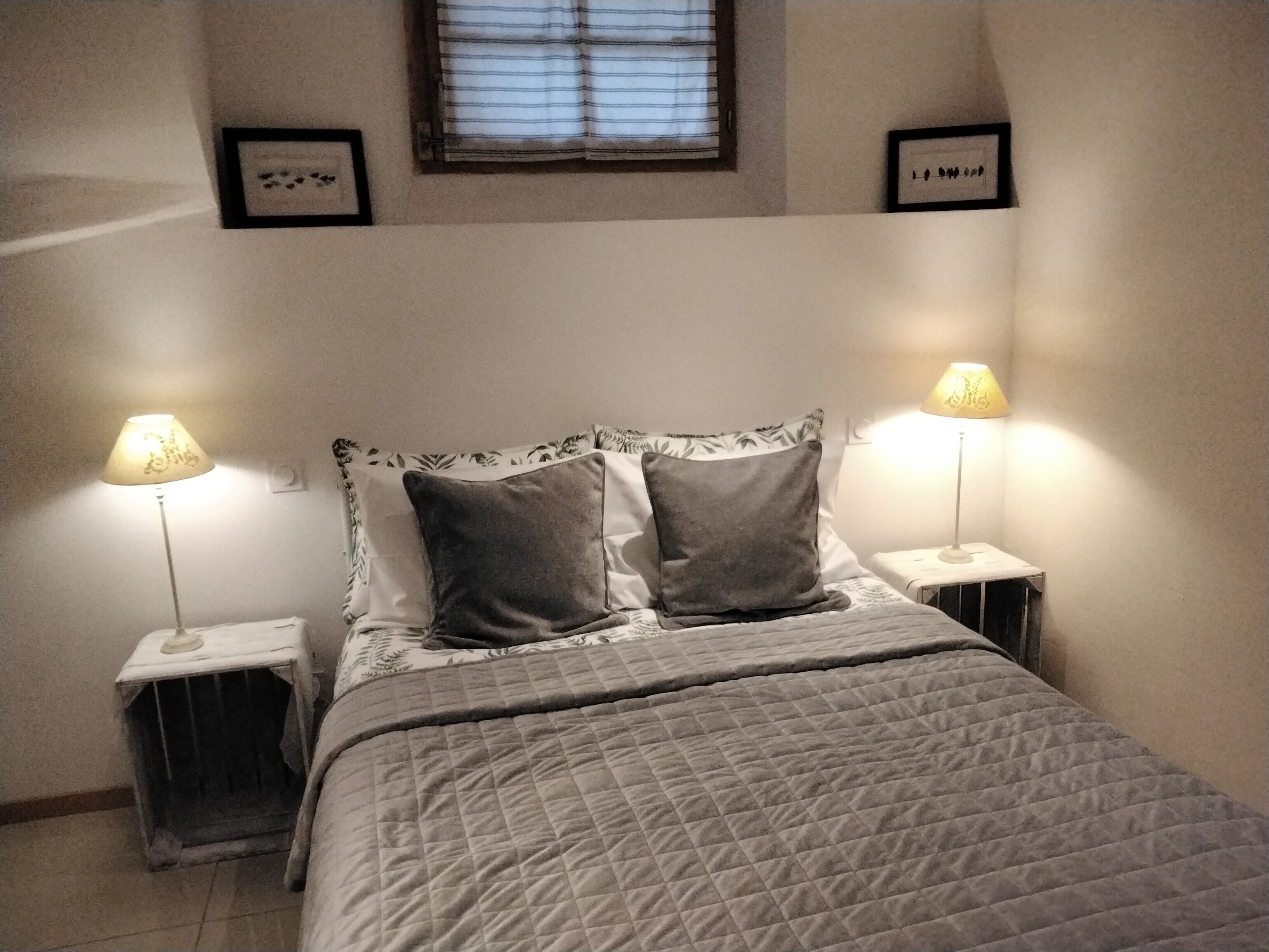 A bedroom with a bed, two bedside tables with lamps, a window with blinds, and framed pictures on the wall.