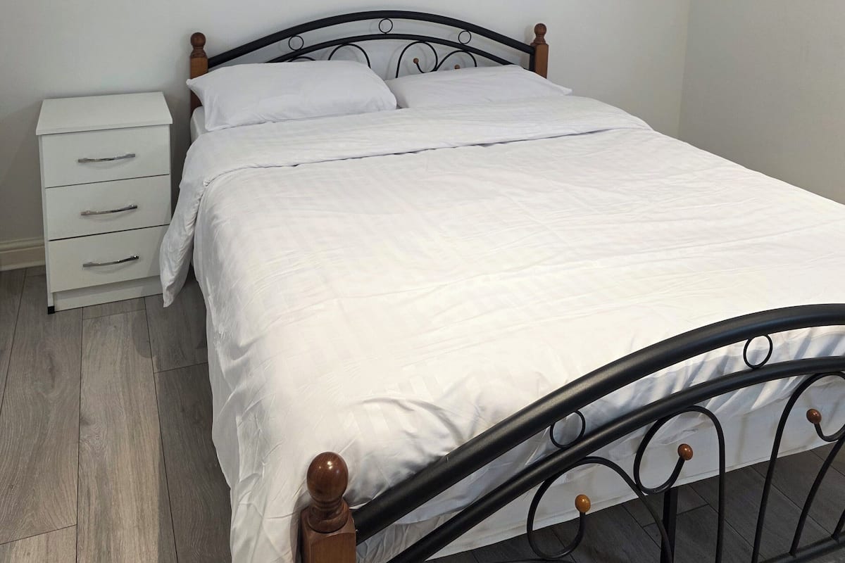 A single bed with a wooden headboard and footboard, a white bedside table with drawers, and a plain white wall.