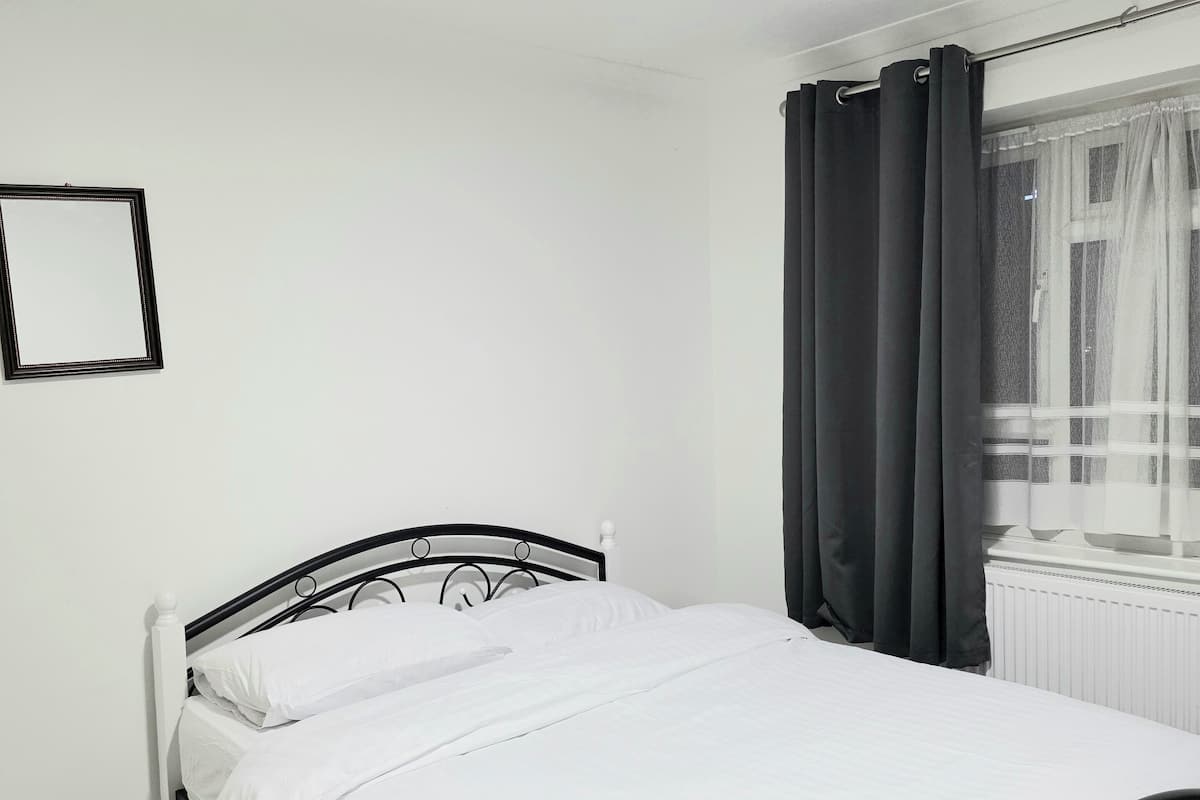 A single bed with white bedding in a room with white walls, a window with dark curtains, and a ceiling light.
