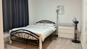 A single bed with a metal frame, a bedside table, a floor lamp, and a standing fan in a room with wooden flooring and a dark curtain. - Baraka House Royal Victoria London by OYO (London)