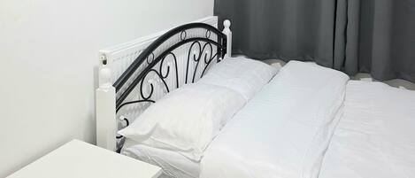 A single bed with white bedding, a white bedside table, and a black metal headboard. Grey curtains are drawn to the side, and a white radiator is visible on the wall.