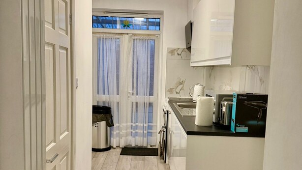 A modern kitchen with white cabinets, a black countertop, and a window with sheer curtains. - Baraka House Royal Victoria London by OYO (London)