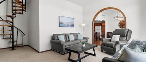 Una sala de estar moderna con una escalera en espiral, un juego de sofá gris, una mesa de centro y un área de comedor visible a través de un arco.