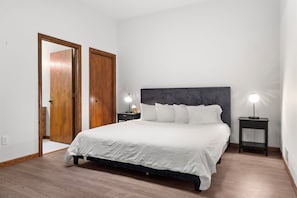 A bedroom with a bed, two bedside tables, a nightstand, and a wooden door. - Loft above the Villager Pub with Round Lake marina sunsets — walk Bridge St + East Park. (Charlevoix)