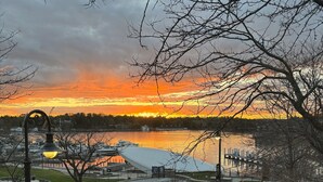 A view of a waterfront area with a sunset, a pedestrian walkway, and a building with a unique roof structure. - Loft above the Villager Pub with Round Lake marina sunsets — walk Bridge St + East Park. (Charlevoix)