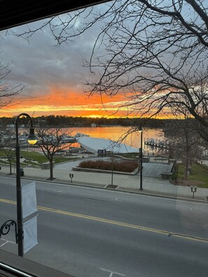 A view of a waterfront area with a sunset, a pedestrian walkway, and a building with a unique roof structure. - Loft above the Villager Pub with Round Lake marina sunsets — walk Bridge St + East Park. (Charlevoix)