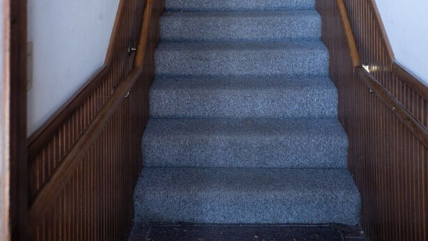 A staircase with wooden handrails and carpeted steps leading to a landing with a light fixture. - Loft above the Villager Pub with Round Lake marina sunsets — walk Bridge St + East Park. (Charlevoix)