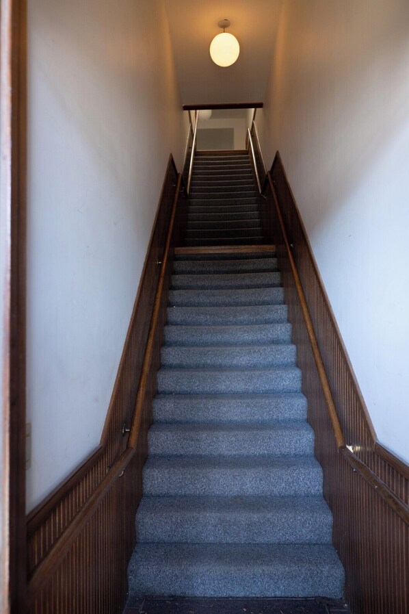 A staircase with wooden handrails and carpeted steps leading to a landing with a light fixture. - Loft above the Villager Pub with Round Lake marina sunsets — walk Bridge St + East Park. (Charlevoix)