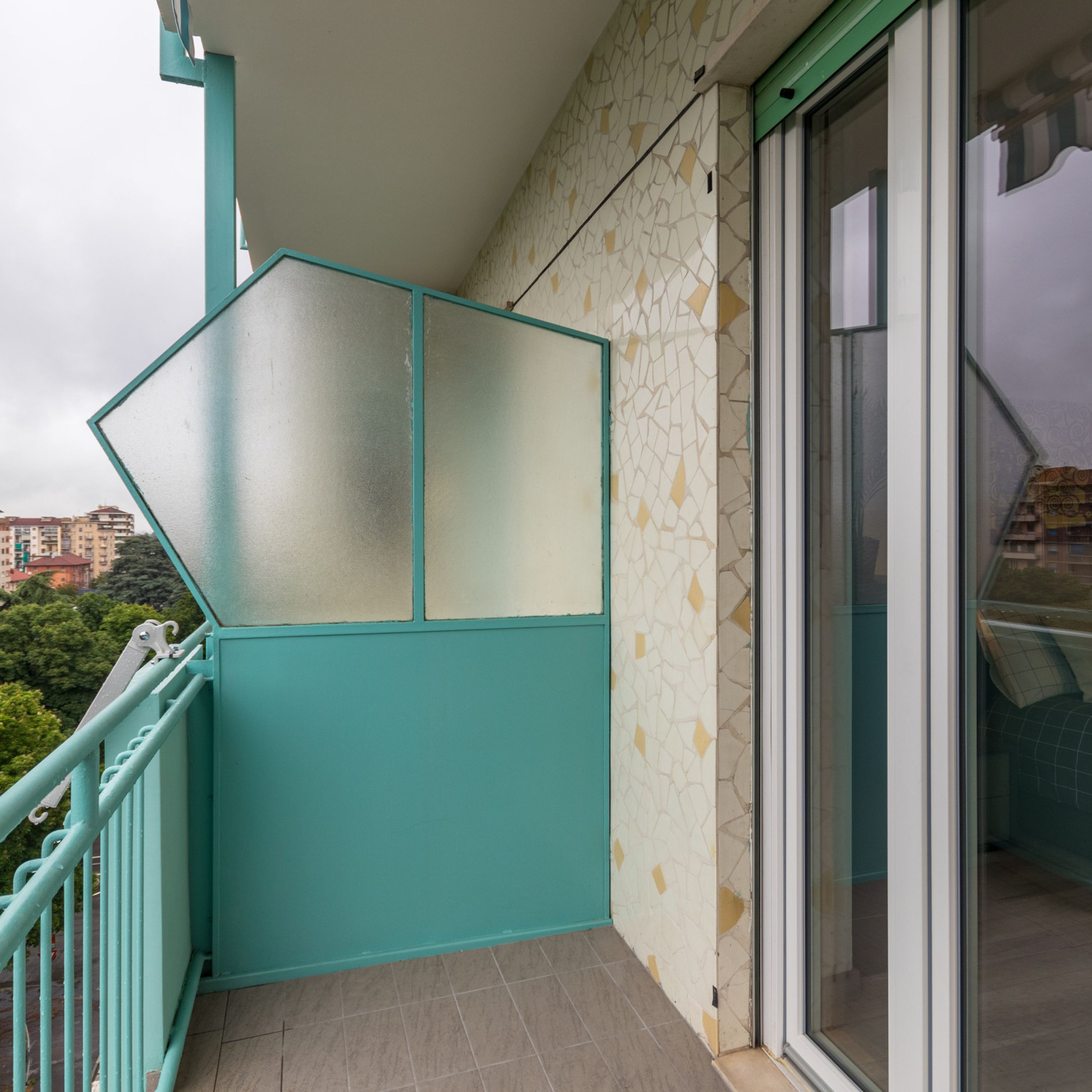 Ein Balkon mit türkisblauem Geländer, einer Glastür und einer Wand mit Muster.