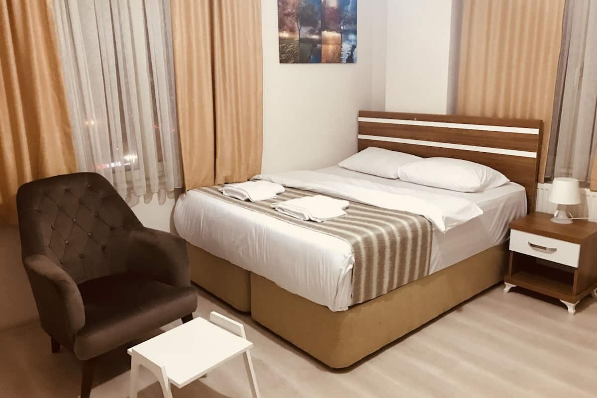 Quarto de hotel bem arrumado, com uma cama, uma cadeira, uma mesinha e uma mesa de cabeceira com abajur.