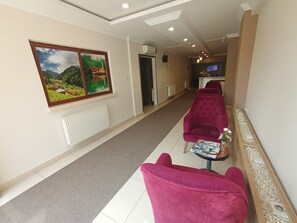 Ein Hotel-Rezeptionsbereich mit einer Rezeption, einer Wartezone mit zwei roten Sesseln, einer Zeitschrift auf einem kleinen Tisch und einem großen Landschaftsbild an der Wand.