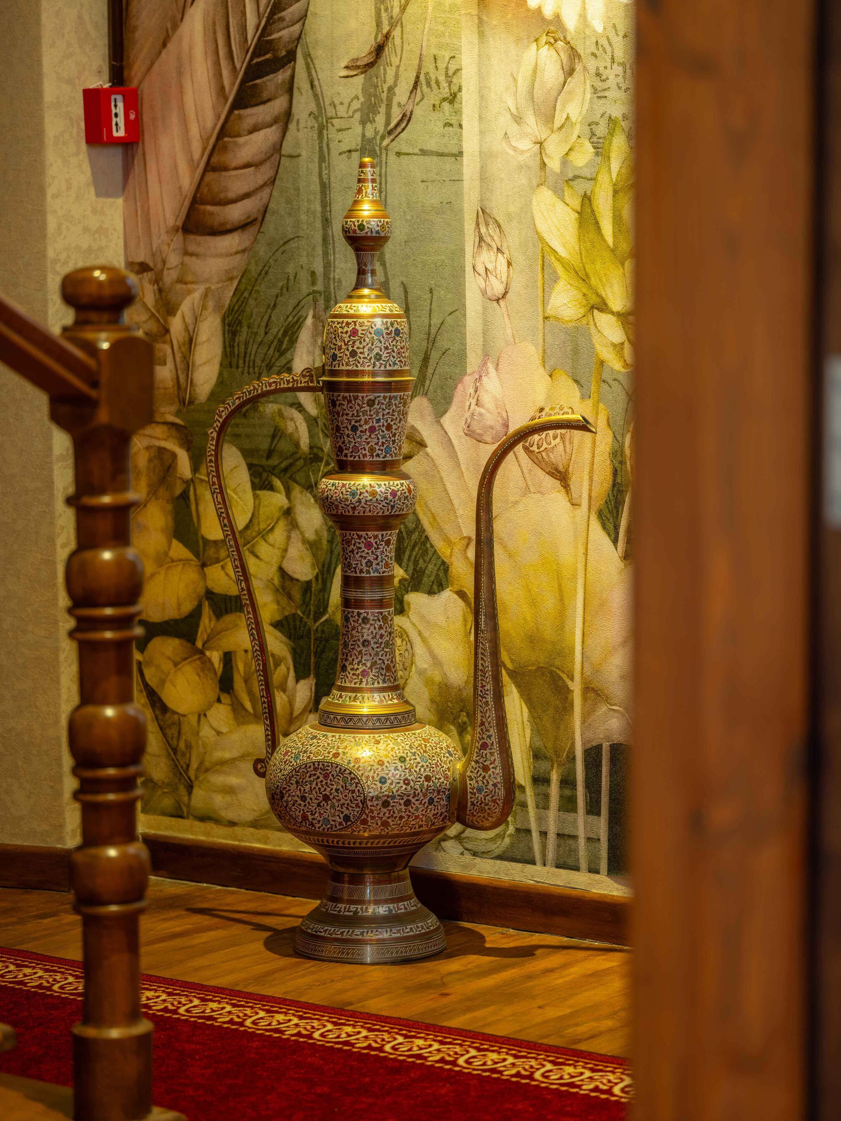 A traditional ornate water vessel with intricate designs, placed on a wooden floor with a red patterned carpet.
