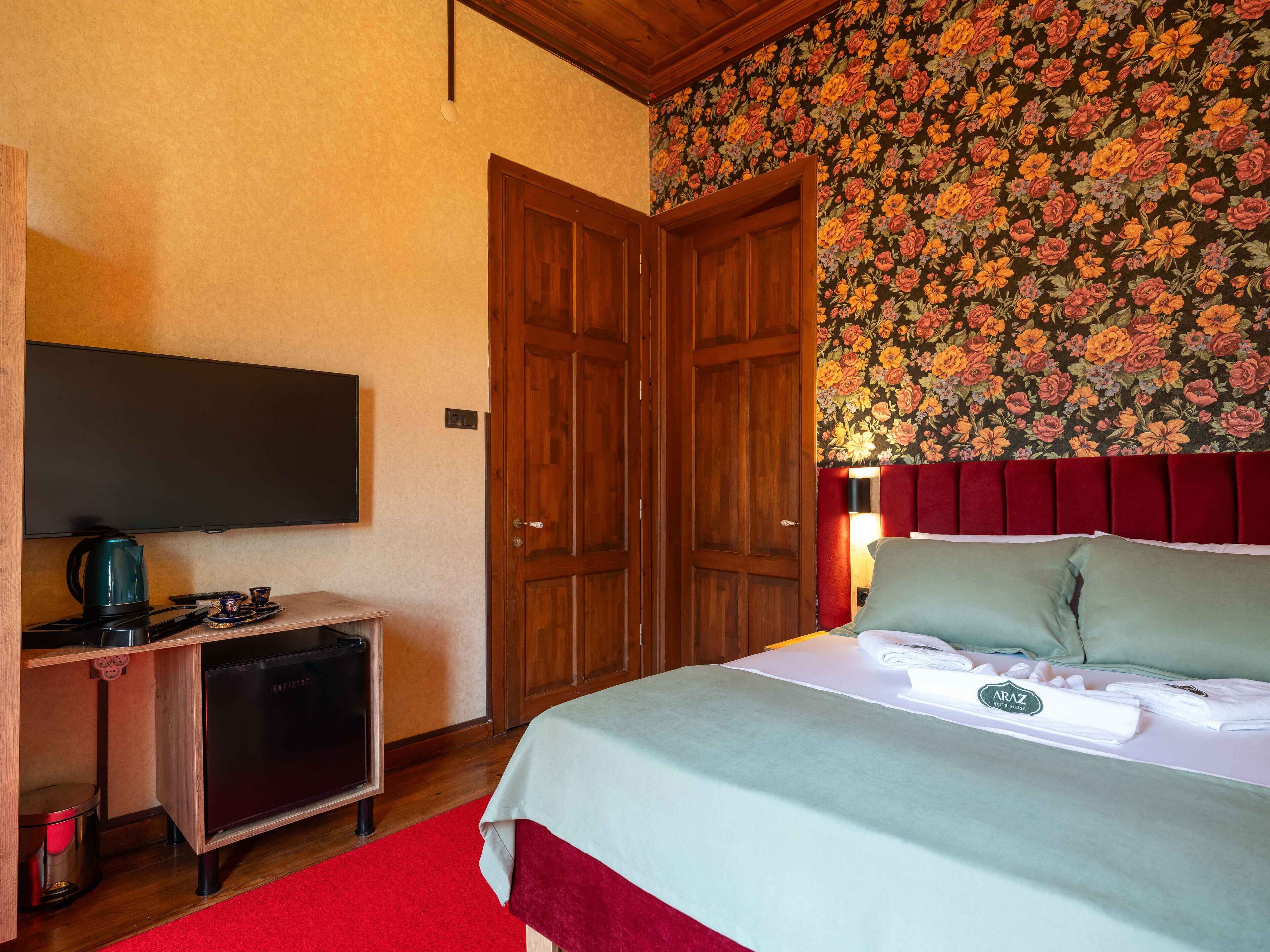 A hotel room with a bed, a television, a desk with a kettle, and a red carpet.