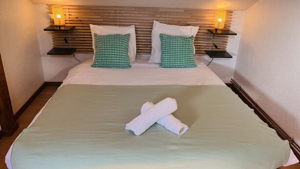 Ein Bett mit einer grünen Decke, zwei Kissen mit Karomuster und einem aufgerollten Handtuch.