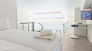 A modern hotel room with a large bed, a desk with a chair, a television, and a balcony with a view of the sea.
