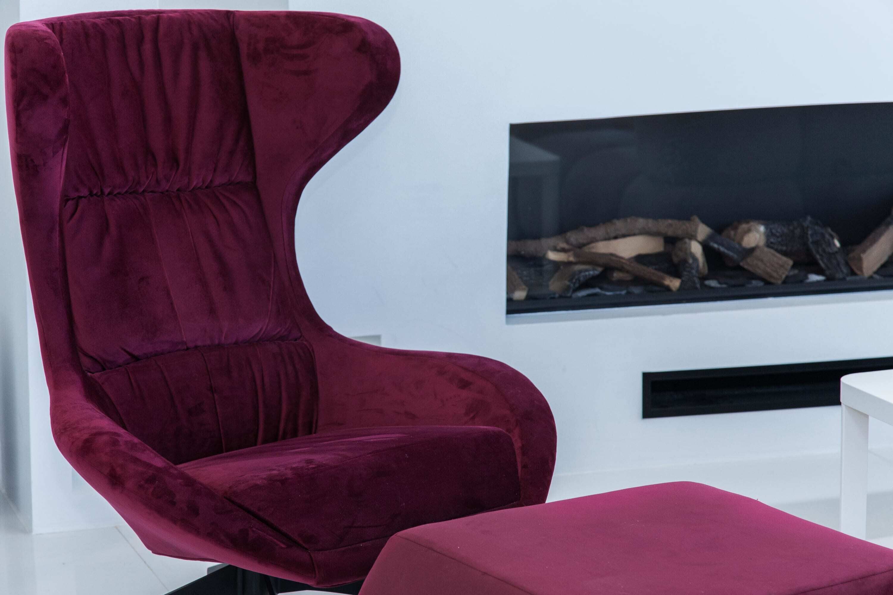 Ein modernes Wohnzimmer mit einem bequemen, rot-samtenen Sessel und Hocker, einem weißen Couchtisch und einem Kamin mit Holzscheiten.