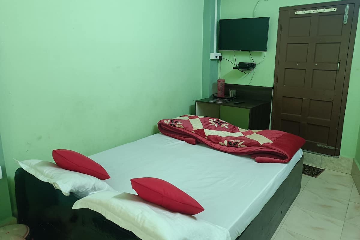 A single bed with a red and white quilt, two red pillows, and a white mattress cover. There is a small table with a TV and a brown door in the background.