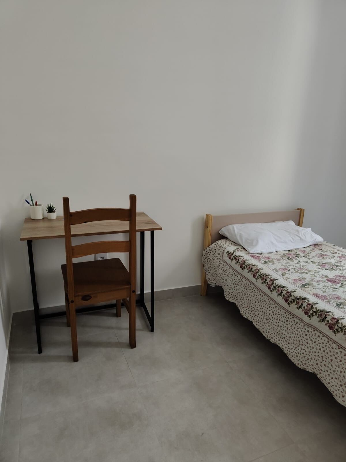 Et lille, pænt møbleret soveværelse med et træskrivebord, en stol og en enkelt seng med blomstret sengetæppe.