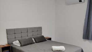 A neatly made bed with a grey headboard and pillows, a bedside table with a book, and a wall-mounted air conditioner.