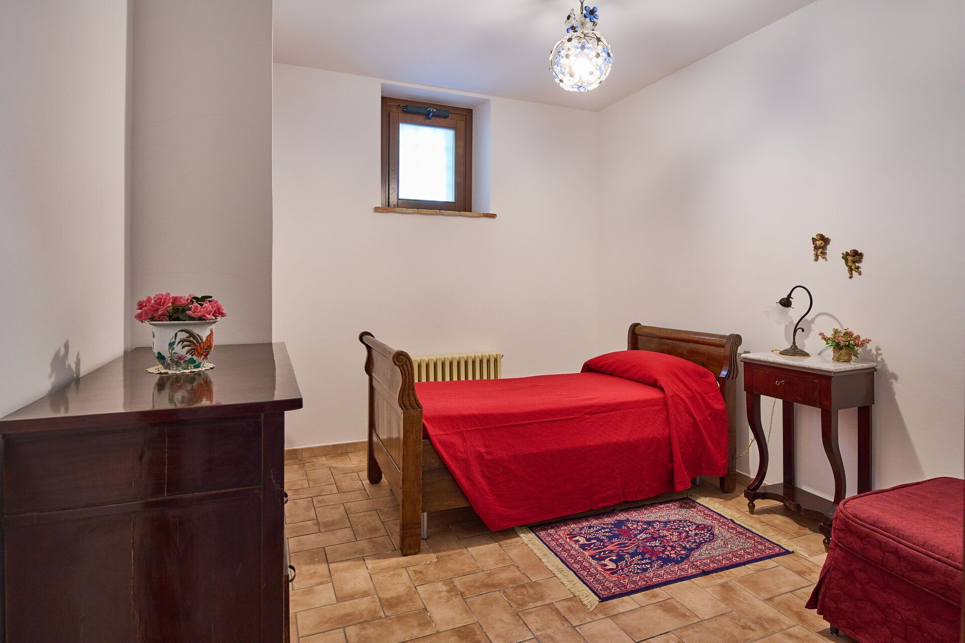 A bedroom with a wooden bed, a red bedspread, a radiator, a small table with a lamp, a vase with flowers, and a window.