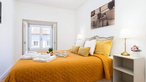 Una habitación de hotel con cama, ventana, mesita de noche con lámpara y un cuadro en la pared.