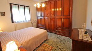 Un dormitorio con cama, armario de madera, radiador, ventana con cortinas y mesita de noche con lámpara.