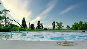 Une piscine avec un escalier en métal, entourée d’une végétation luxuriante et de grands arbres, sous un ciel partiellement nuageux.