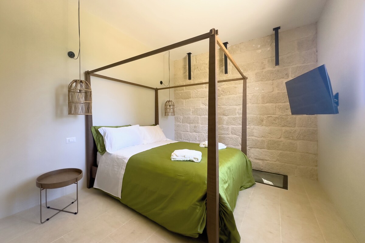 Een hemelbed met een groene sprei en witte kussens, een klein rond tafeltje, en een stenen muur op de achtergrond.