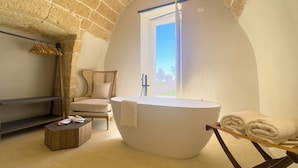 Une pièce spacieuse avec une grande baignoire ovale, une chaise en bois, une petite table et une vue sur l’extérieur par une fenêtre.