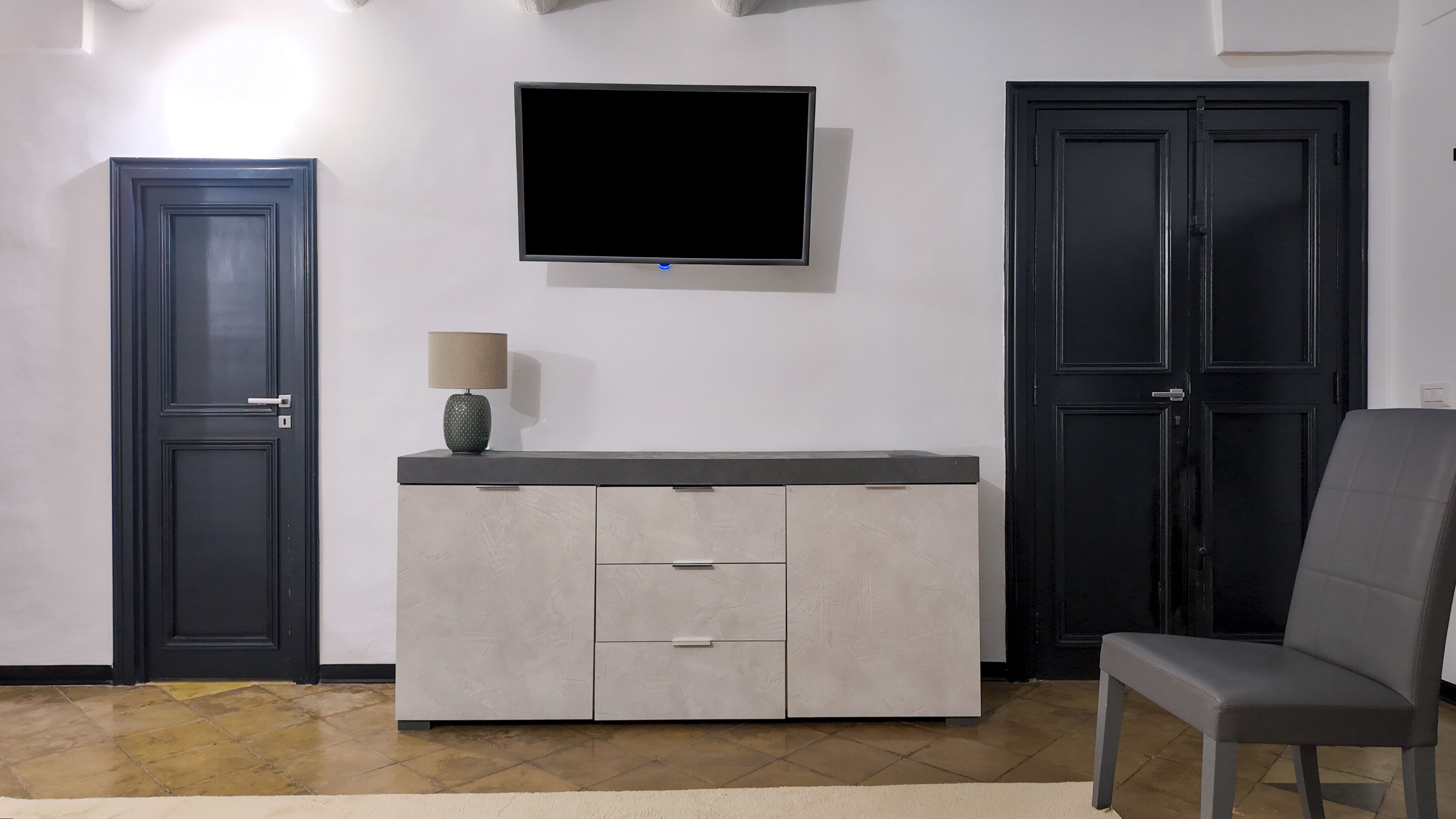 Una habitación con televisión montada en la pared, un aparador con cajones y una silla.