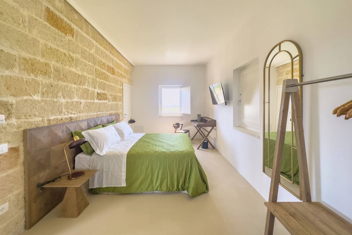 Une chambre avec un mur en briques, un lit avec une literie verte, un bureau, une chaise, un miroir et une fenêtre.