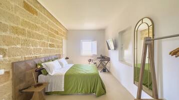 A bedroom with a brick wall, a bed with green bedding, a desk, a chair, a mirror, and a window.