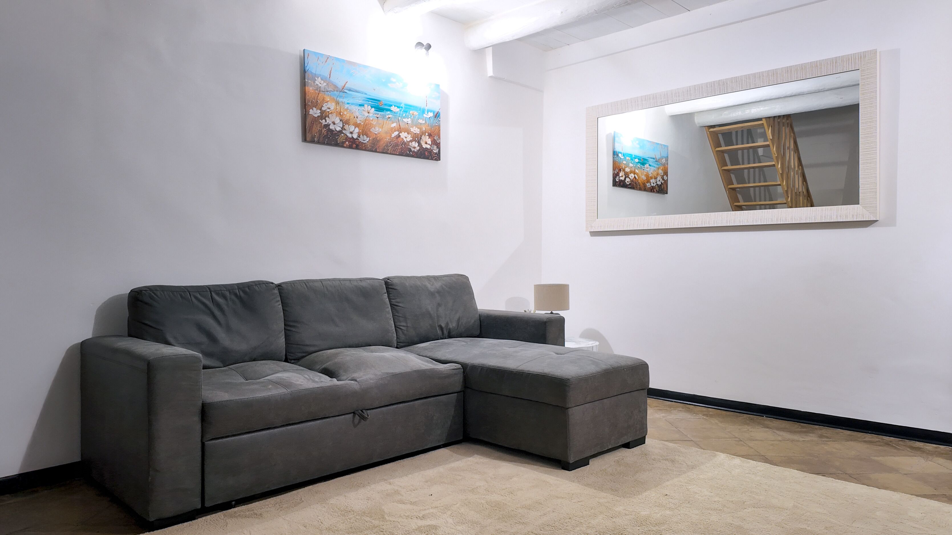 Un canapé d’angle gris dans une pièce aux murs blancs, avec un grand miroir et deux tableaux.