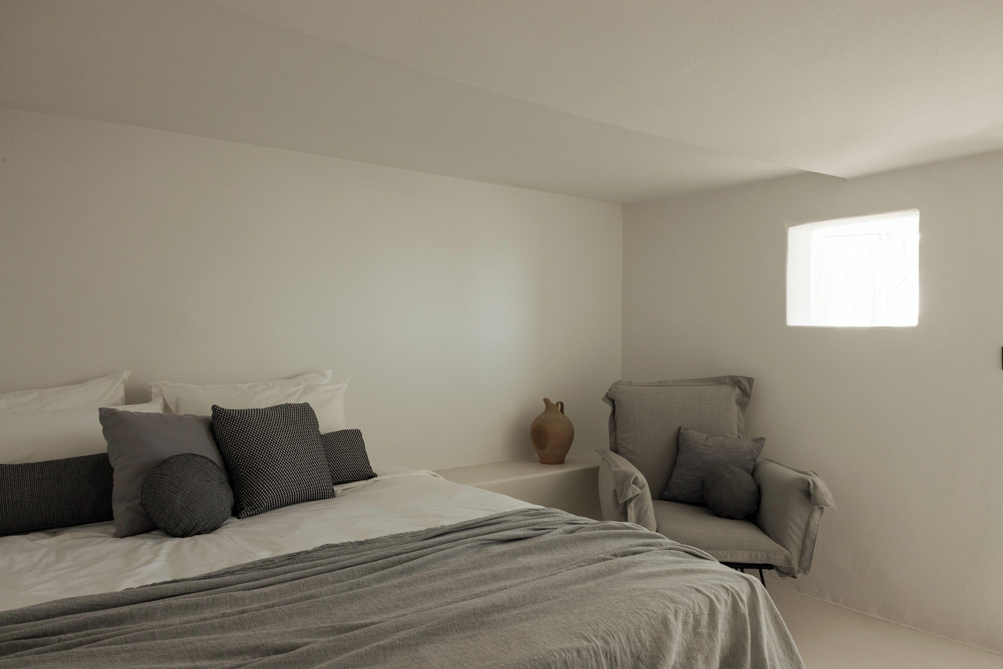 A minimalist bedroom with a large bed, a chair, and a small window.