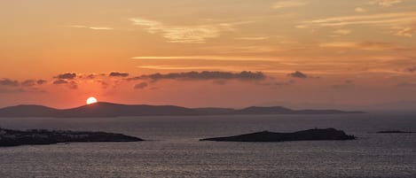 Un pueblo costero al atardecer con el sol poniéndose sobre el mar, edificios a lo largo de la orilla y colinas distantes.