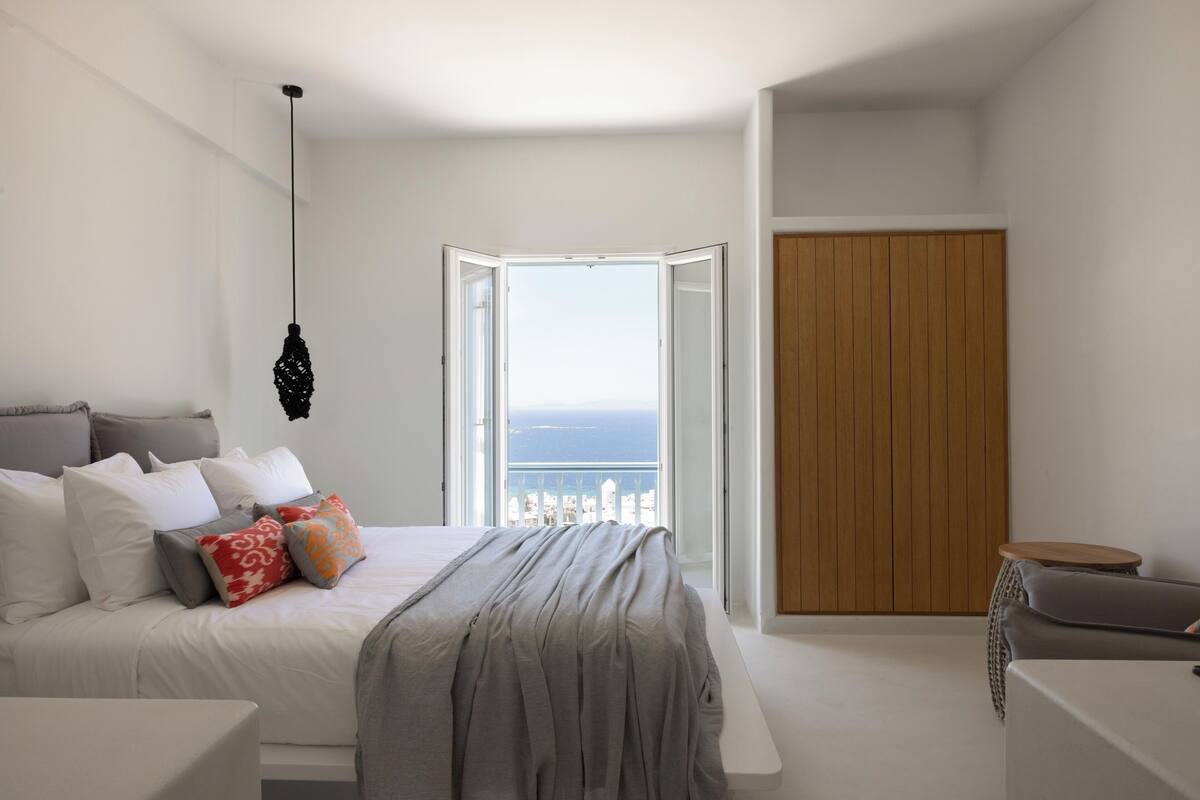 Un dormitorio moderno con cama, lámpara colgante, puerta de madera y vista al mar por una ventana abierta.