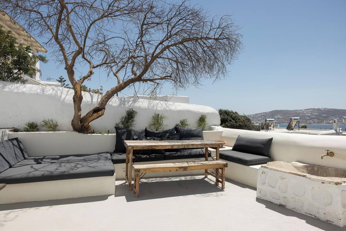 Un espacio al aire libre con luz solar, con una mesa y bancos de madera, rodeado de paredes blancas y vistas al mar.