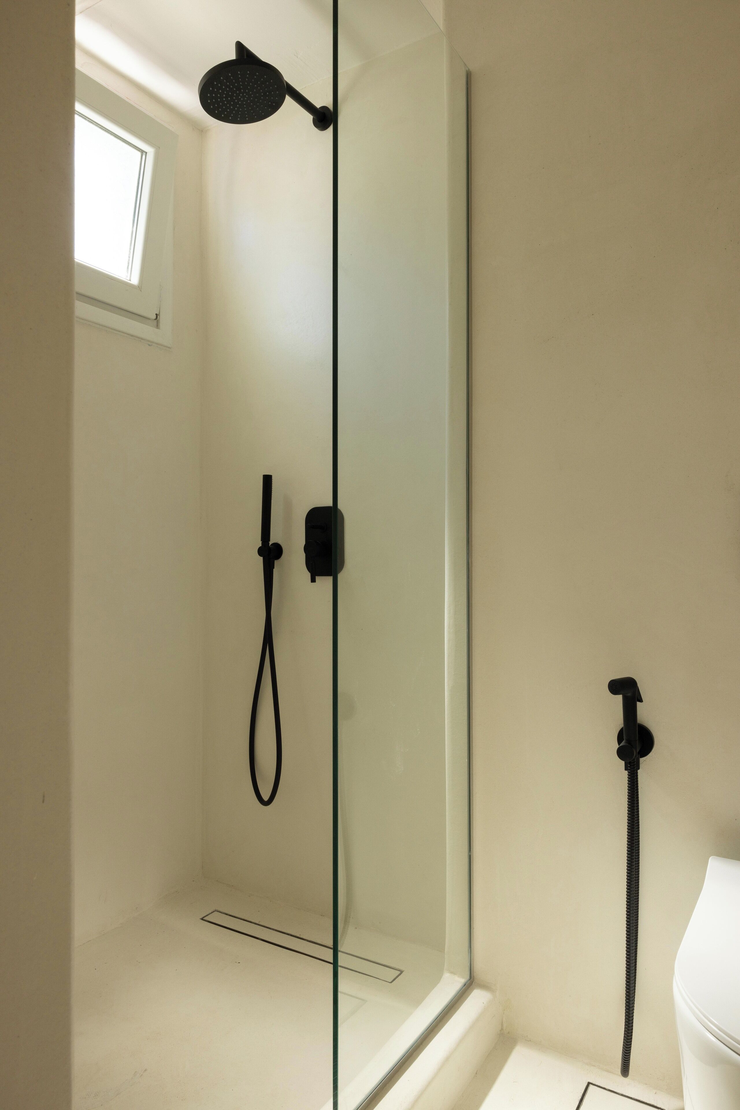 A shower with a handheld showerhead and a wall-mounted faucet in a bathroom with a white toilet and a small window.