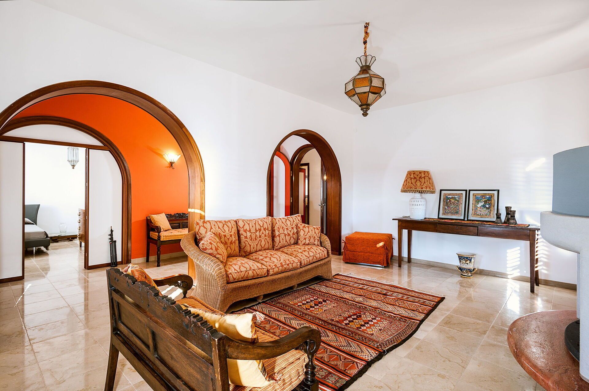 Amplio salón con un sofá grande, un banco de madera y un tapete estampado. Hay una consola con objetos decorativos y una lámpara. La habitación tiene puertas abovedadas y un candelabro.