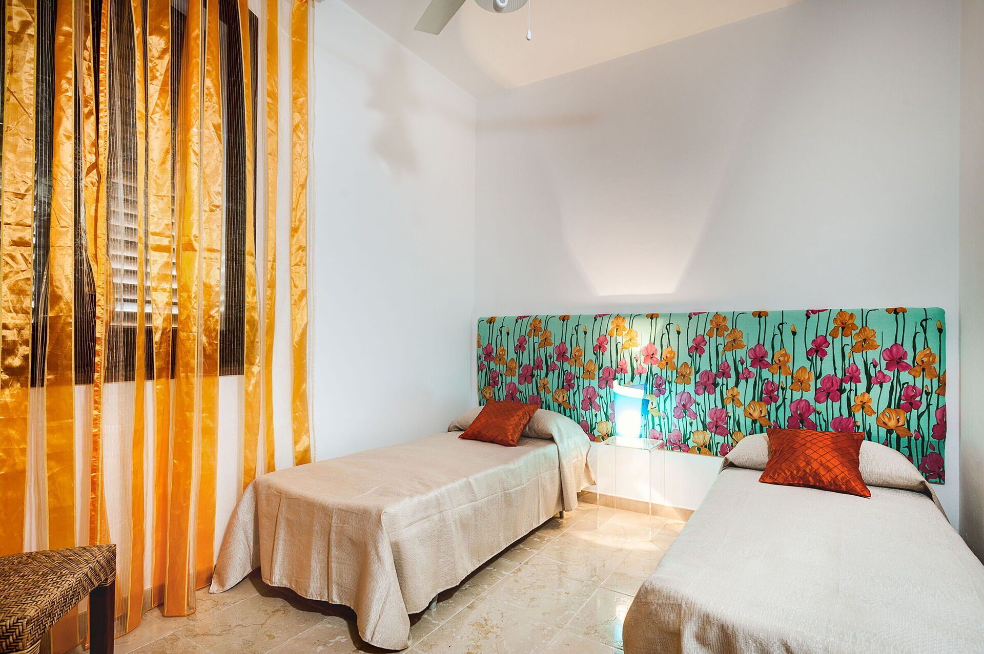 A hotel room with two single beds, a large window with curtains, and a colorful floral headboard.