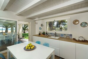 Eine moderne Küche mit Blick auf den Strand, eine Essecke mit weißem Tisch und eine Küchenzeile mit Spüle und dekorativen Tellern.