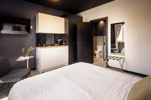 Una habitación de hotel moderna con una cama grande, un escritorio pequeño con una planta, un sofá, un armario y un baño visible a través de una puerta abierta.