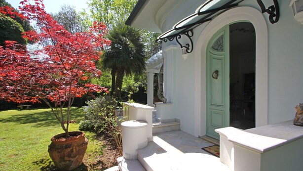 A white building with a green door, a porch with white steps, and a red tree in the garden.