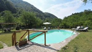 Una piscina con sillas de descanso y una mesa, rodeada por un solárium de piedra y abundante vegetación.