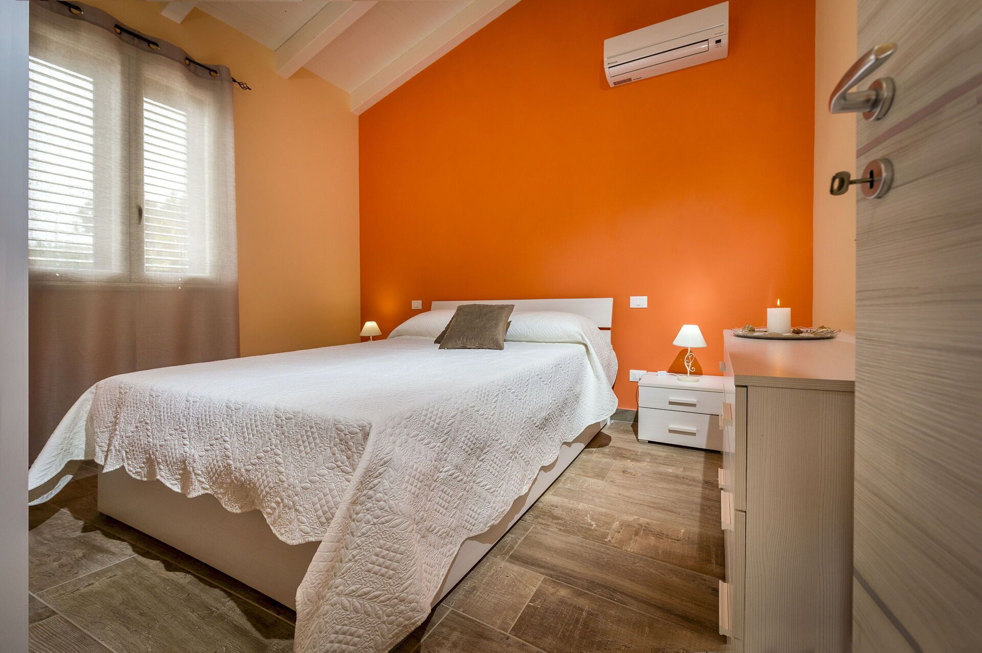 Een slaapkamer met een oranje wand, een wit bed en een nachtkastje met een lamp.
