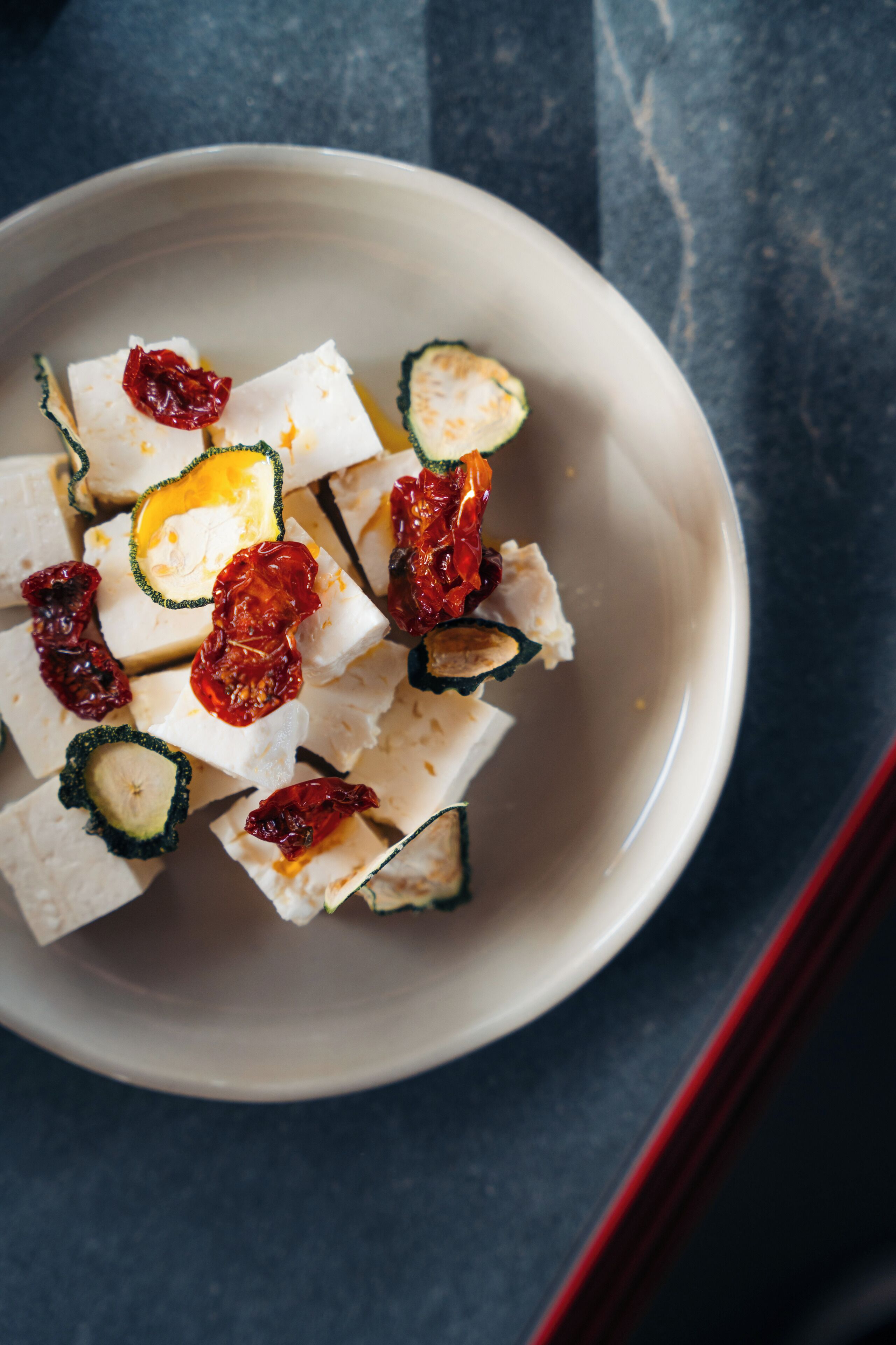 Un tazón de cubos de tofu con tomates secos al sol sobre una superficie oscura.