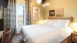 Une chambre avec un grand lit, un bureau et une chaise. Il y a une fenêtre avec des rideaux, un tableau au mur et un appareil de climatisation.