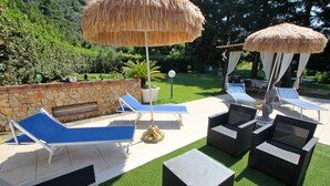 Una terrazza esterna con poltrone relax, un muro in pietra e un ombrellone di paglia.